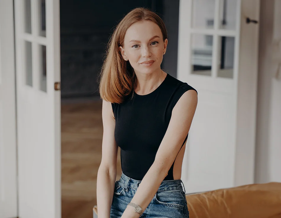 Woman in a black sleeveless top and jeans sitting on a couch, smiling softly at the camera in a cozy indoor setting - Arm Lift in Houston TX