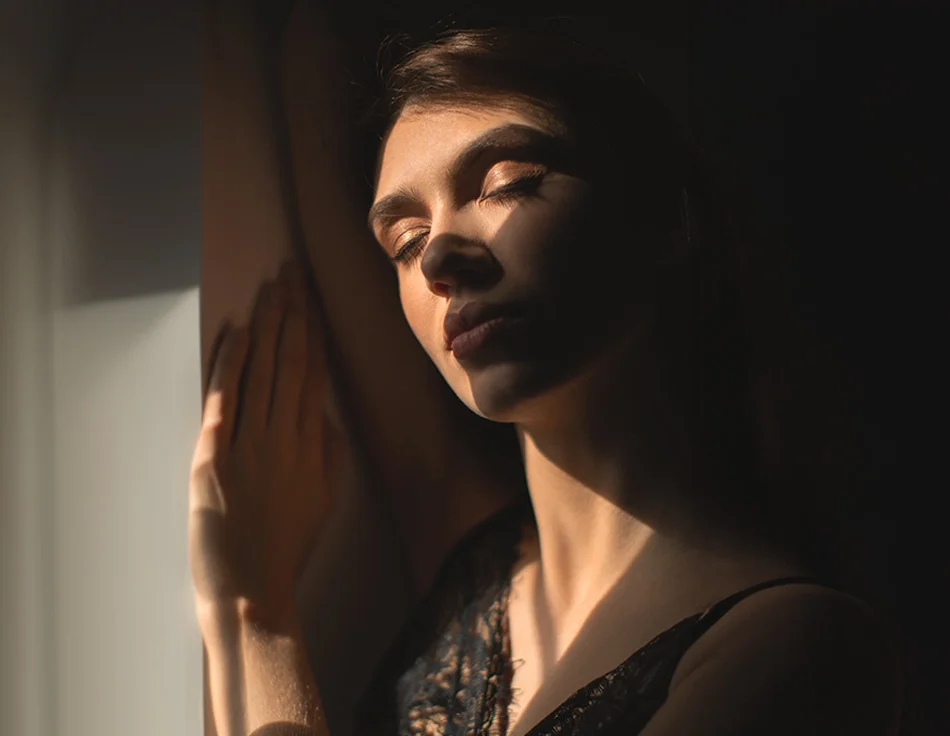 A woman standing in a dark room, with her eyes closed, her face partially illuminated by sunlight - Co2 Laser Resurfacing in Houston, TX