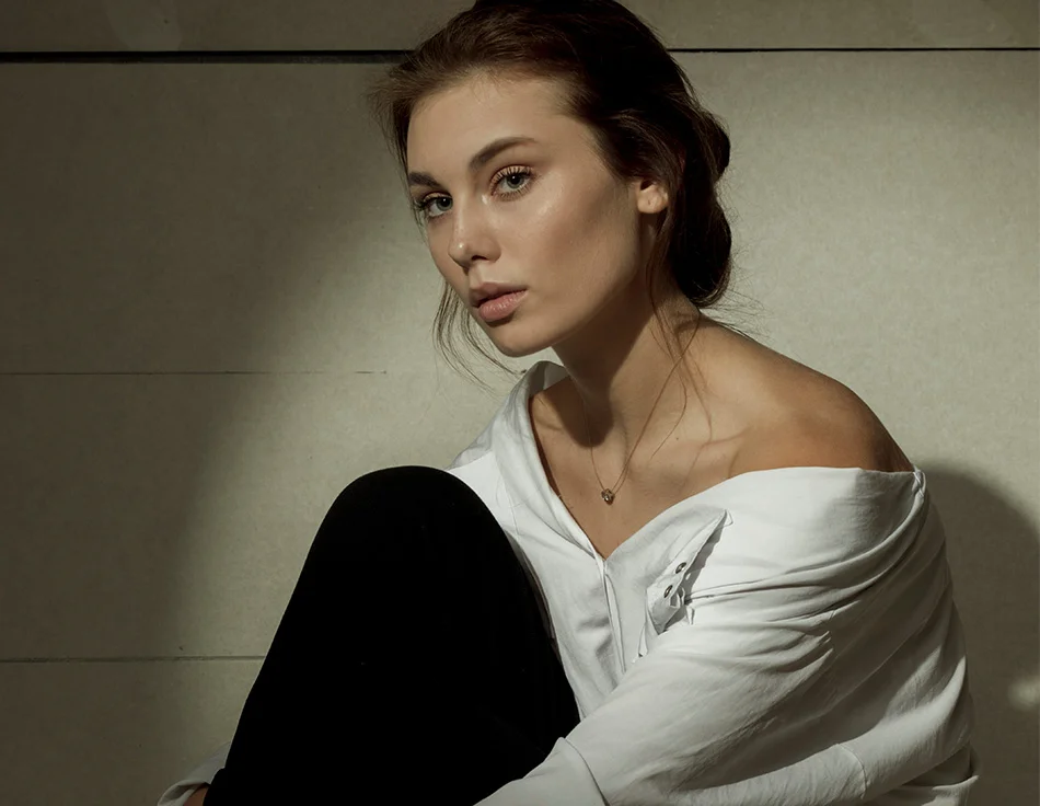 Woman with light brown hair wearing a white off-shoulder blouse, sitting on the floor with a calm and introspective expression - Dermal Fillers in Houston, TX