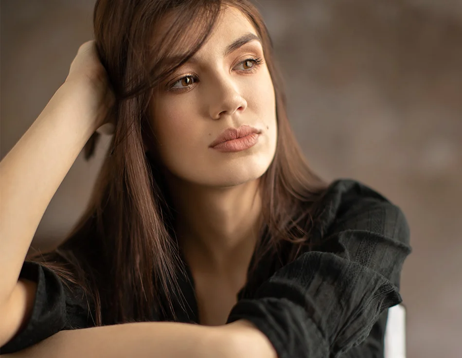 Woman with long brown hair, dressed in a black shirt, resting her head on her hand while looking contemplatively off-camera - Fraxel Laser Procedure in Houston, TX